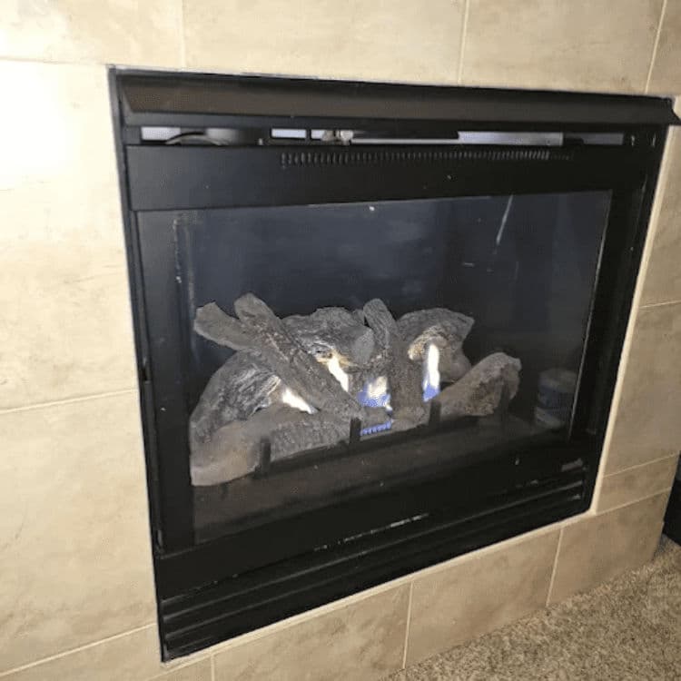 Gas fireplace with glowing logs and flames, framed by a tiled wall. Cozy indoor ambiance.
