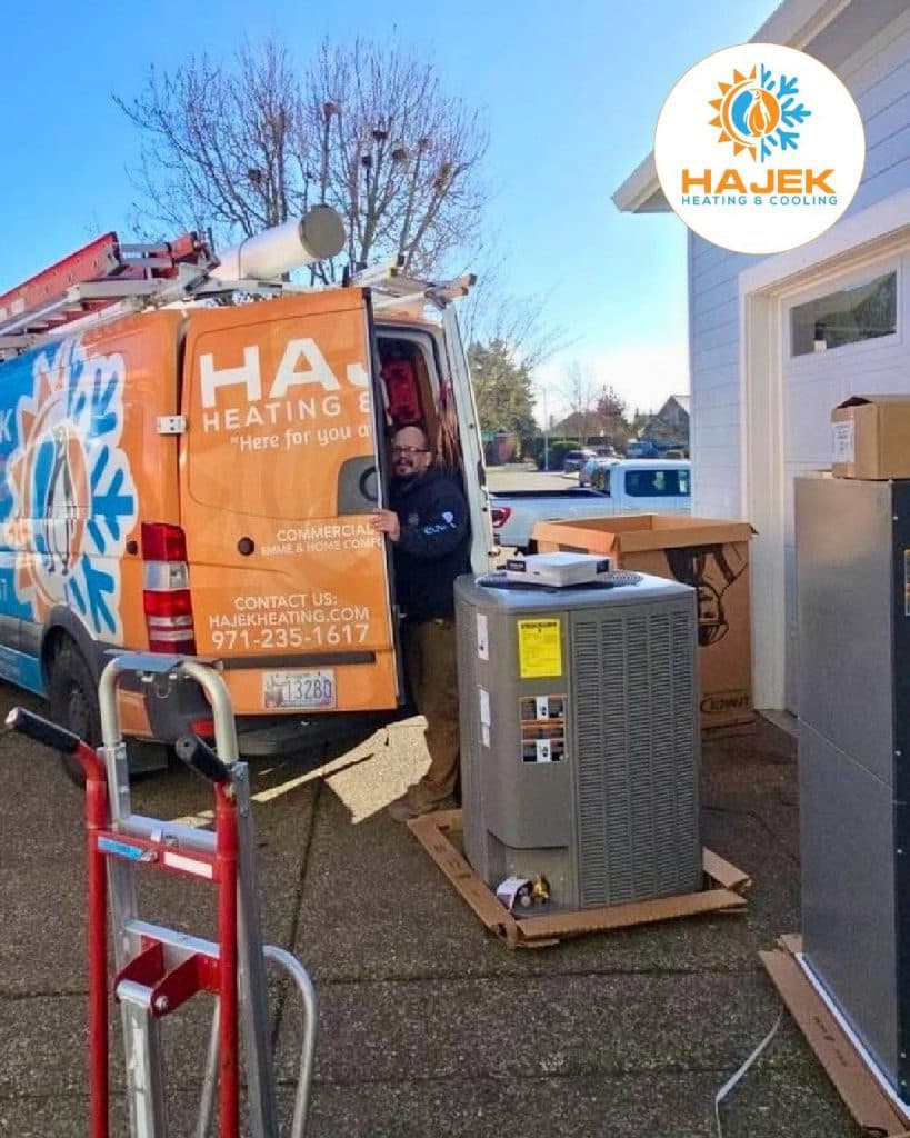HVAC technician unloading equipment from a van marked 'Hajek Heating & Cooling' outdoors.