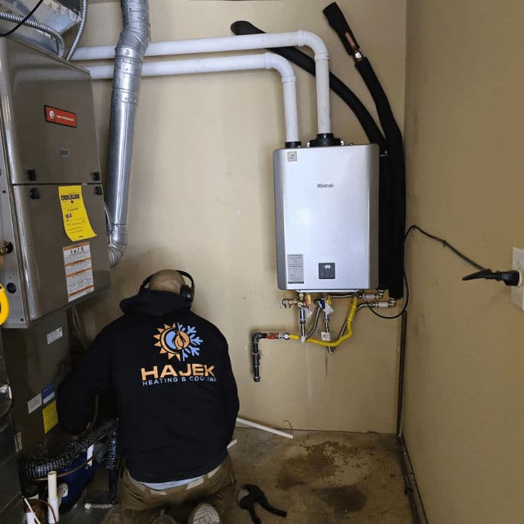Technician servicing a water heater in a residential utility room. Hajek Heating & Cooling logo visible.