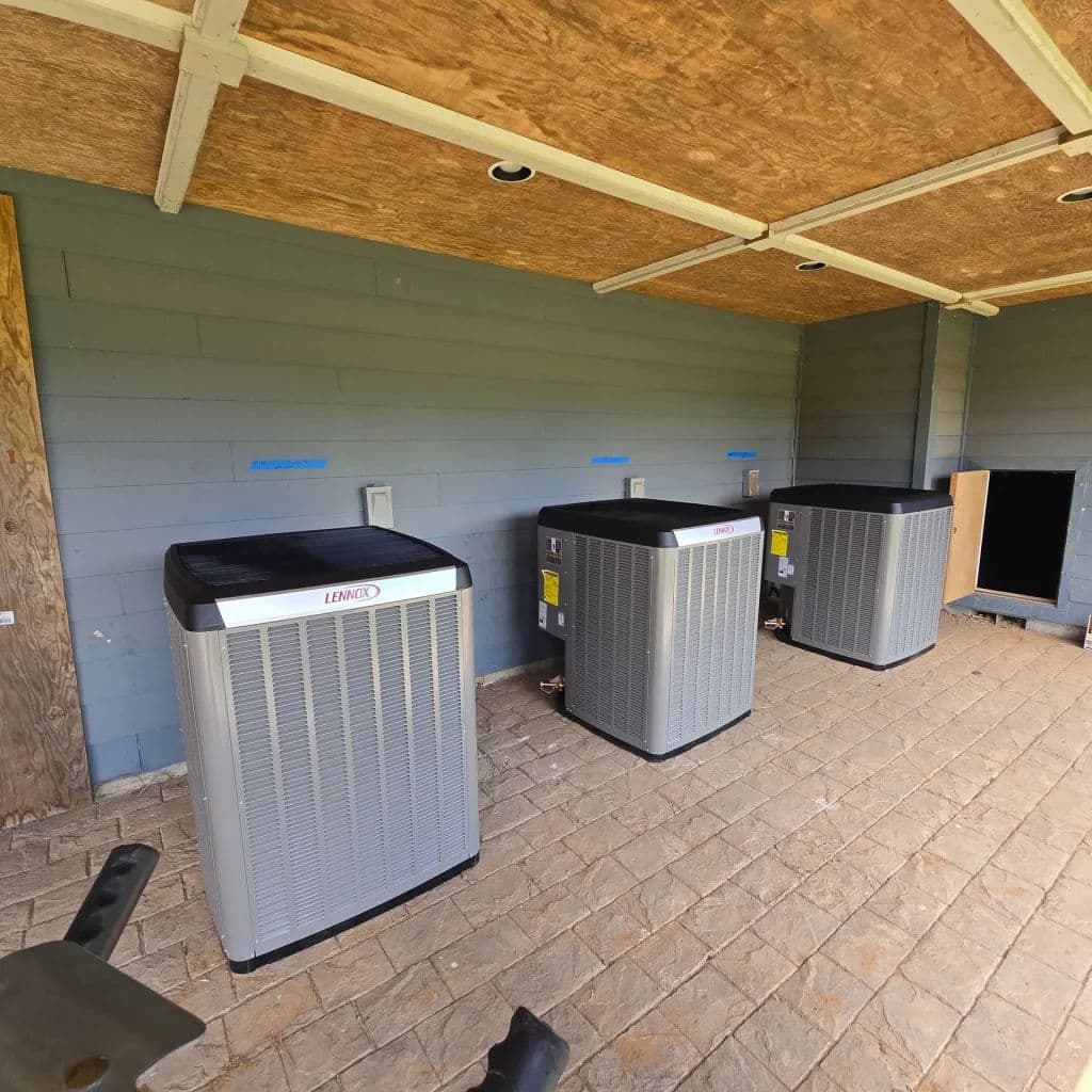 Three outdoor air conditioning units mounted against a wooden wall with stone flooring.
