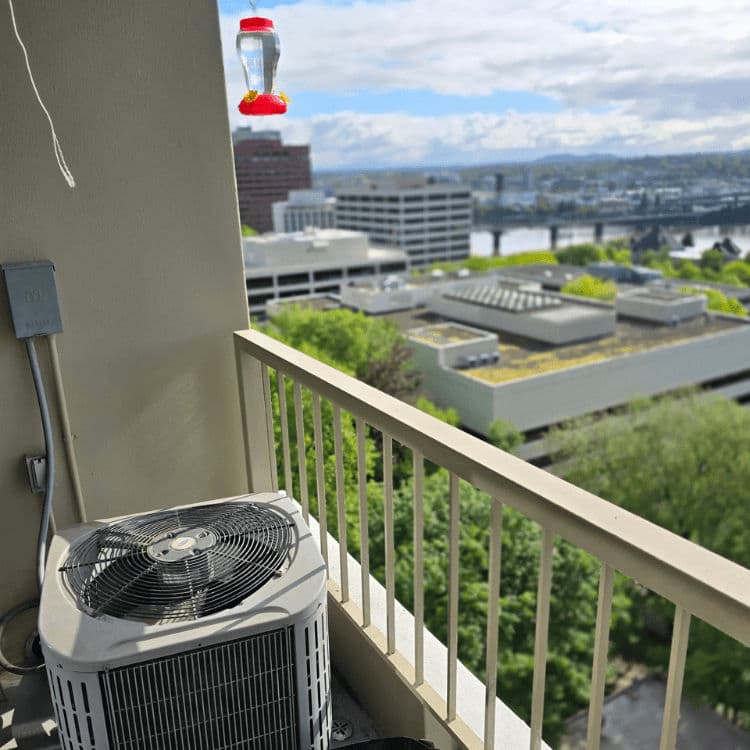 Air conditioning unit on a balcony with a hummingbird feeder and city view in the background.
