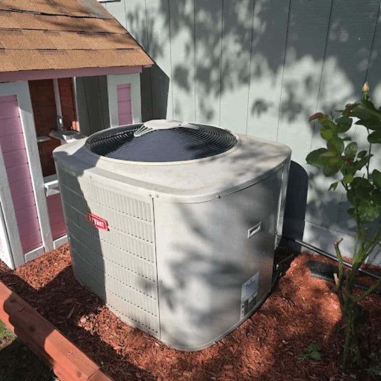 Air conditioning unit installed beside a small wooden playhouse in a garden setting.