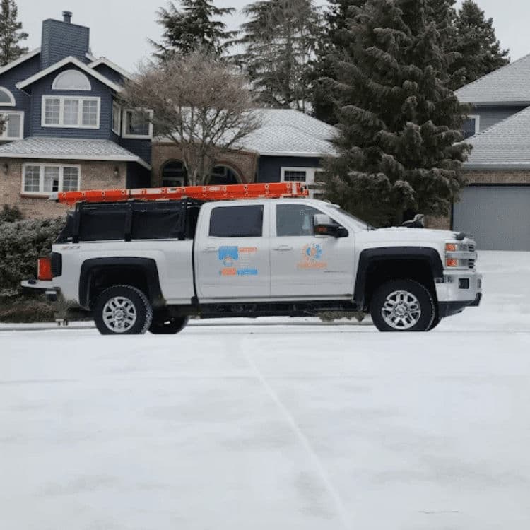 White pickup truck with a ladder rack parked on snow-covered driveway near residential home.