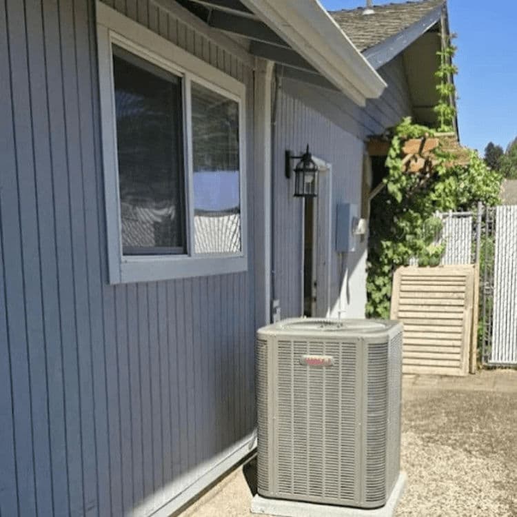 Air conditioning unit next to a gray house with a window and nearby greenery.