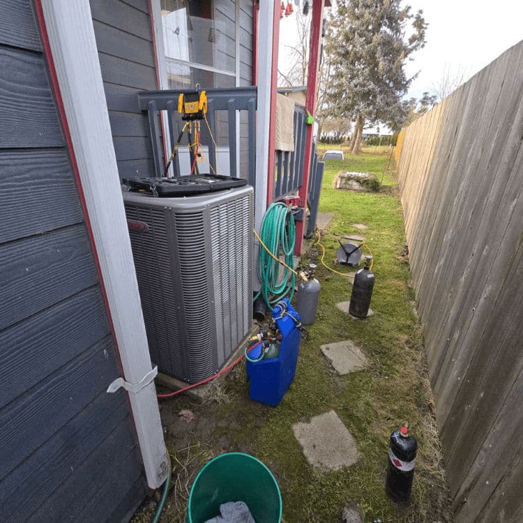 Air conditioning unit beside a house with hoses and maintenance equipment in a grassy area.