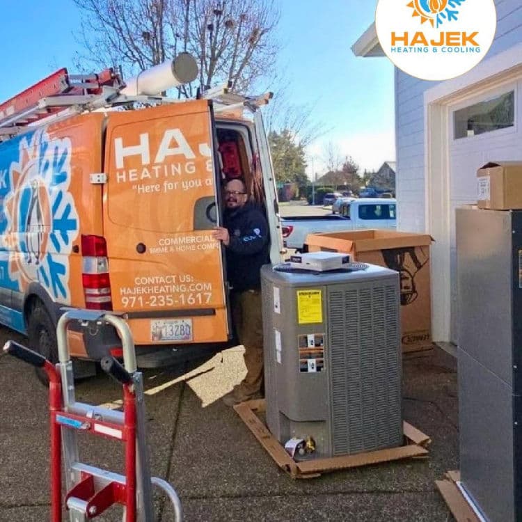Technician unloading HVAC system from Hajek Heating and Cooling van outside a building.